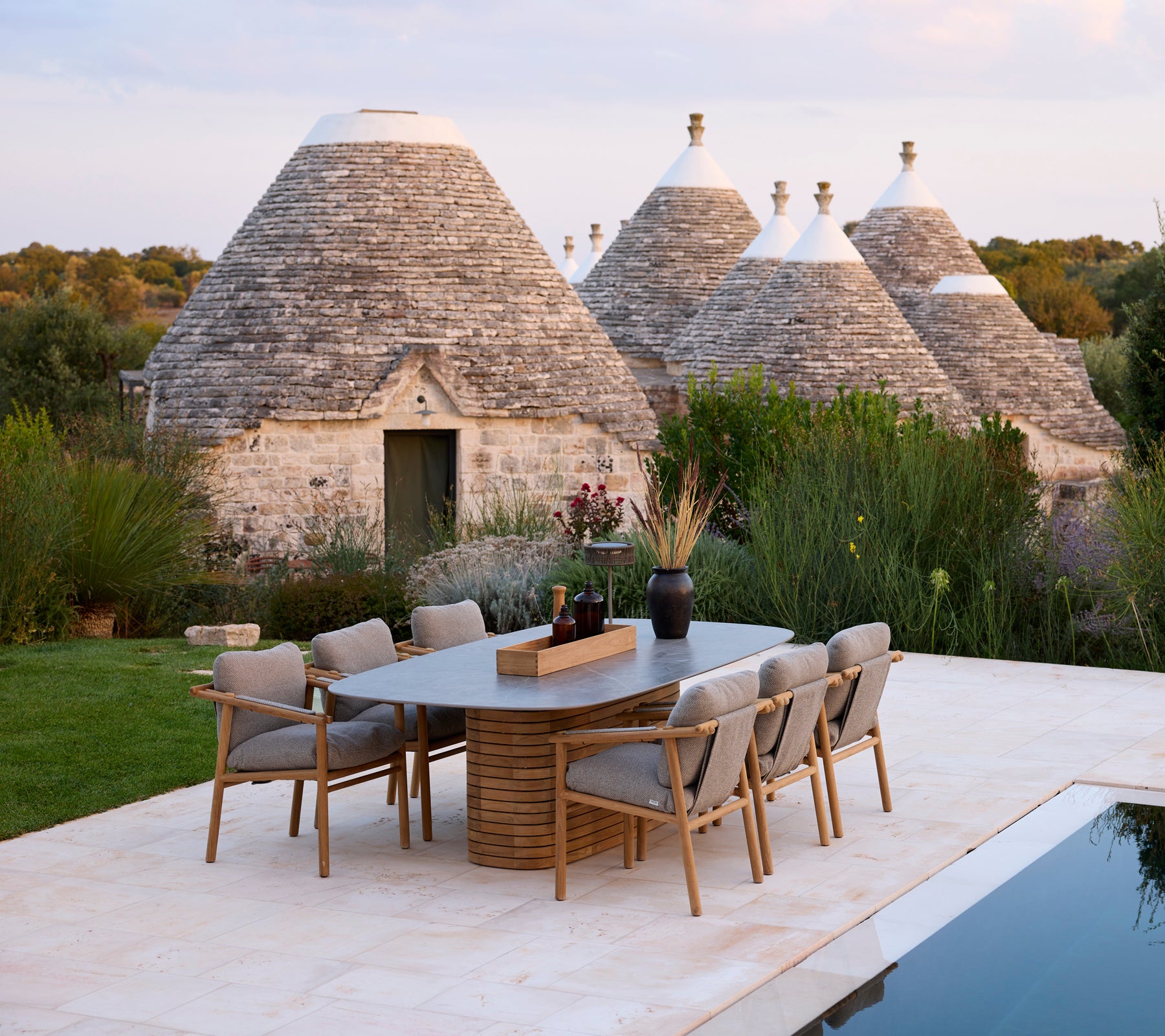 Grå spisebordsstole omkring et rektangulært bord nær traditionelle trullo-strukturer.