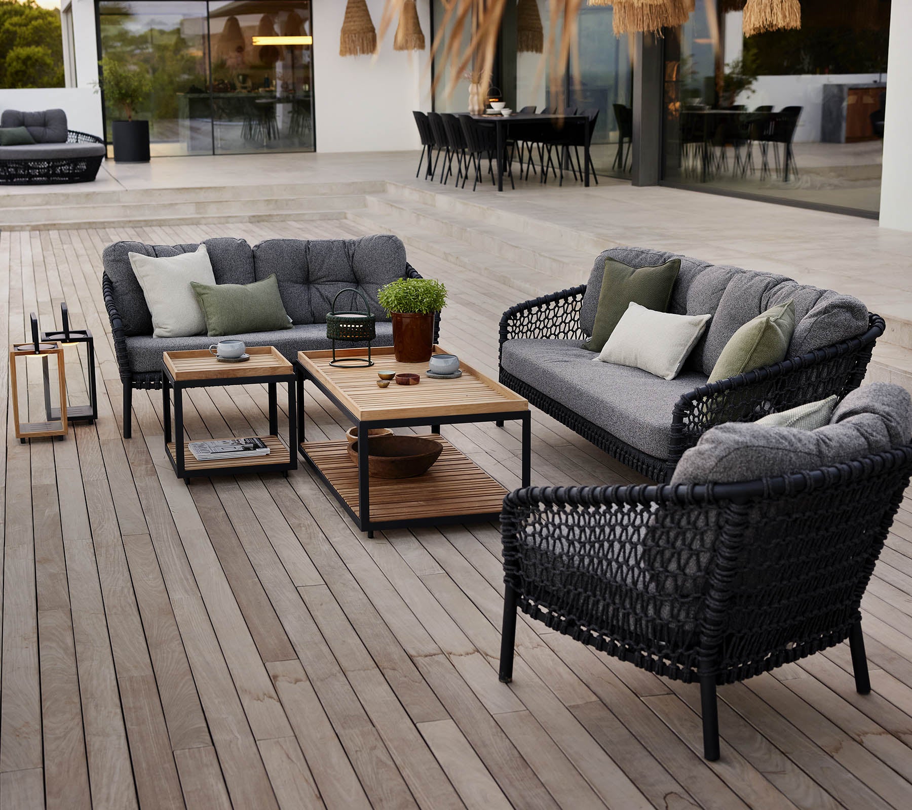 Stilfuld udendørs loungeområde med sort rattanmøbler, grå puder, et trækaffebord og potteplanter på en træterrasse.