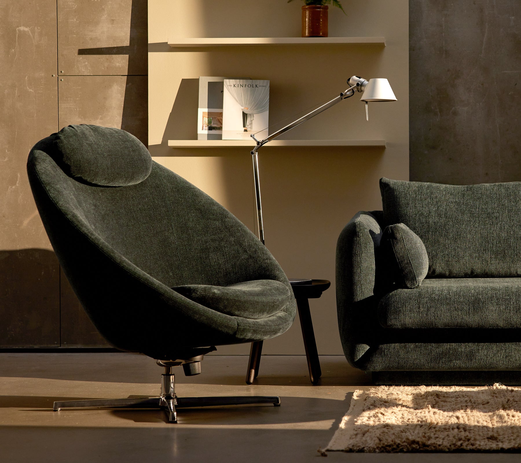 Pace stol i mørkegrøn chenille ved siden af en Bloom sofa, komplementeret af en slank lampe og minimalistisk hyldeindretning.