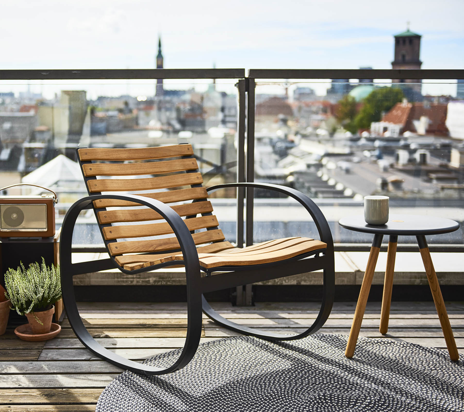En stilfuld gyngestol ved siden af et lille sidebord, sat op mod en panoramisk udsigt over byen, smykket med en plante og et tæppe.