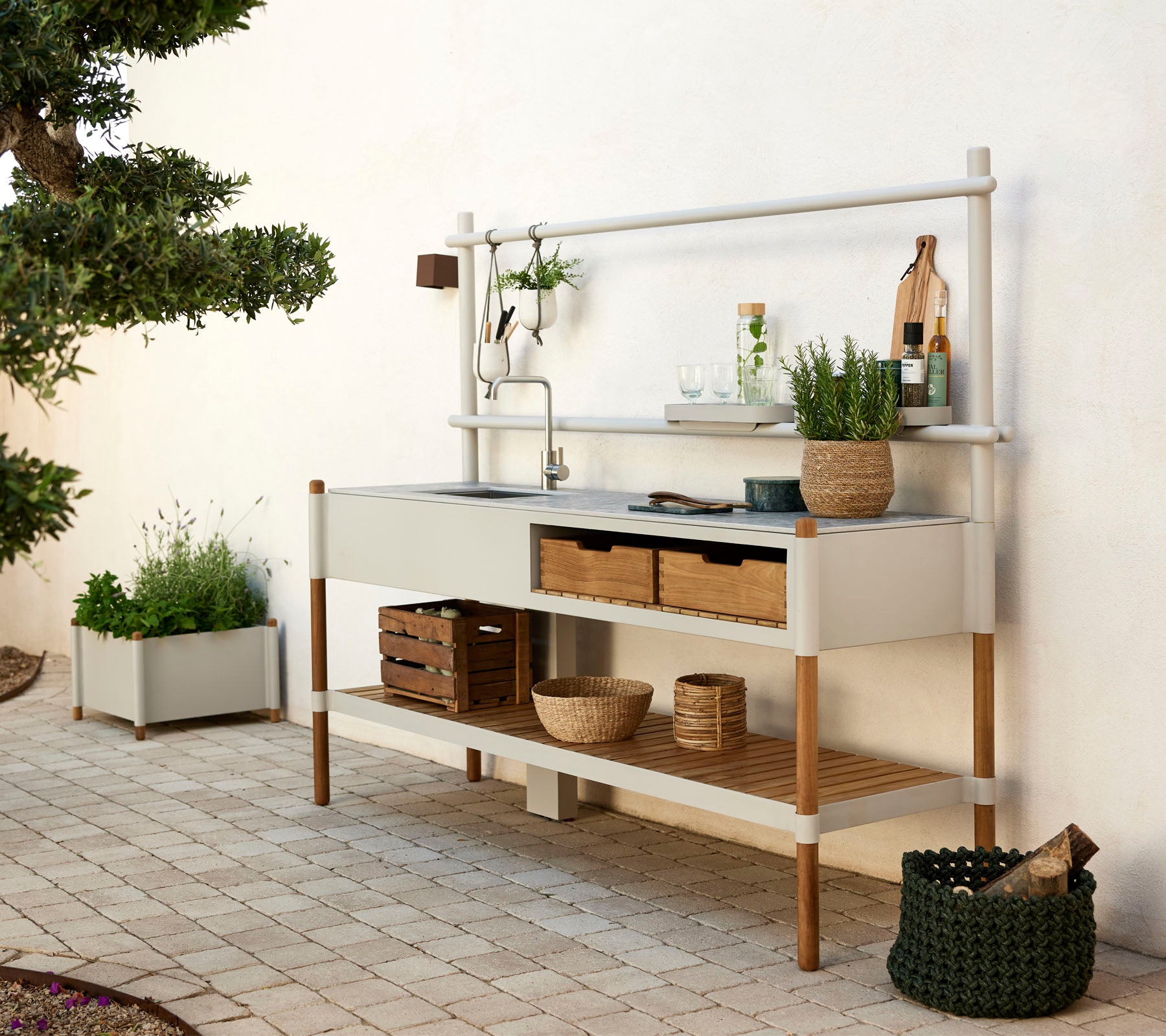 Moderne køkkenenhed med en metalvask, hylder af træ og dekorative planter i stilfulde beholdere. Rent design med naturlige elementer.
