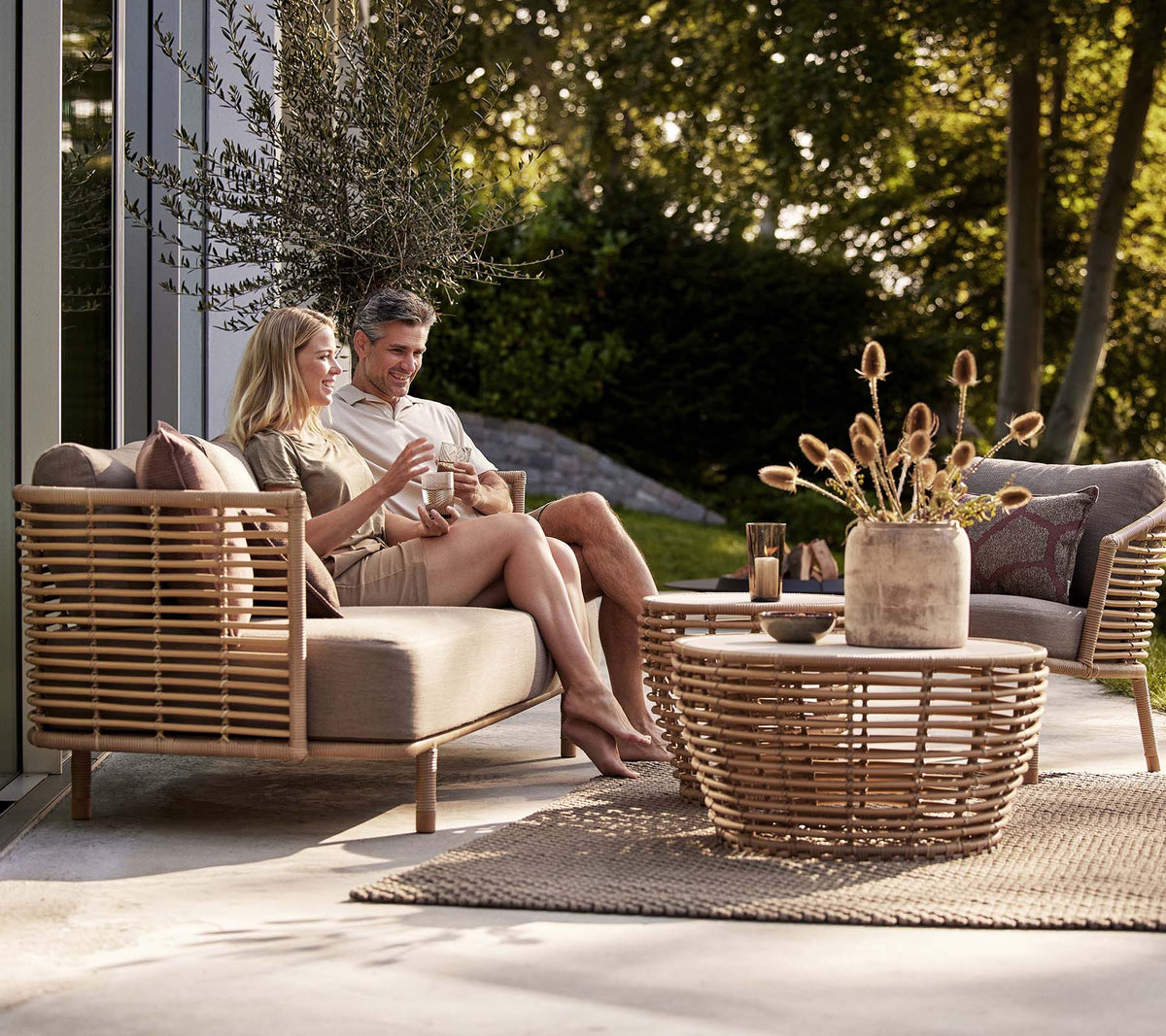 Et par nyder drinks på en Sense-sofa og lounge stol, ledsaget af et stilfuldt kurvekaffebord og en dekorativ vase.