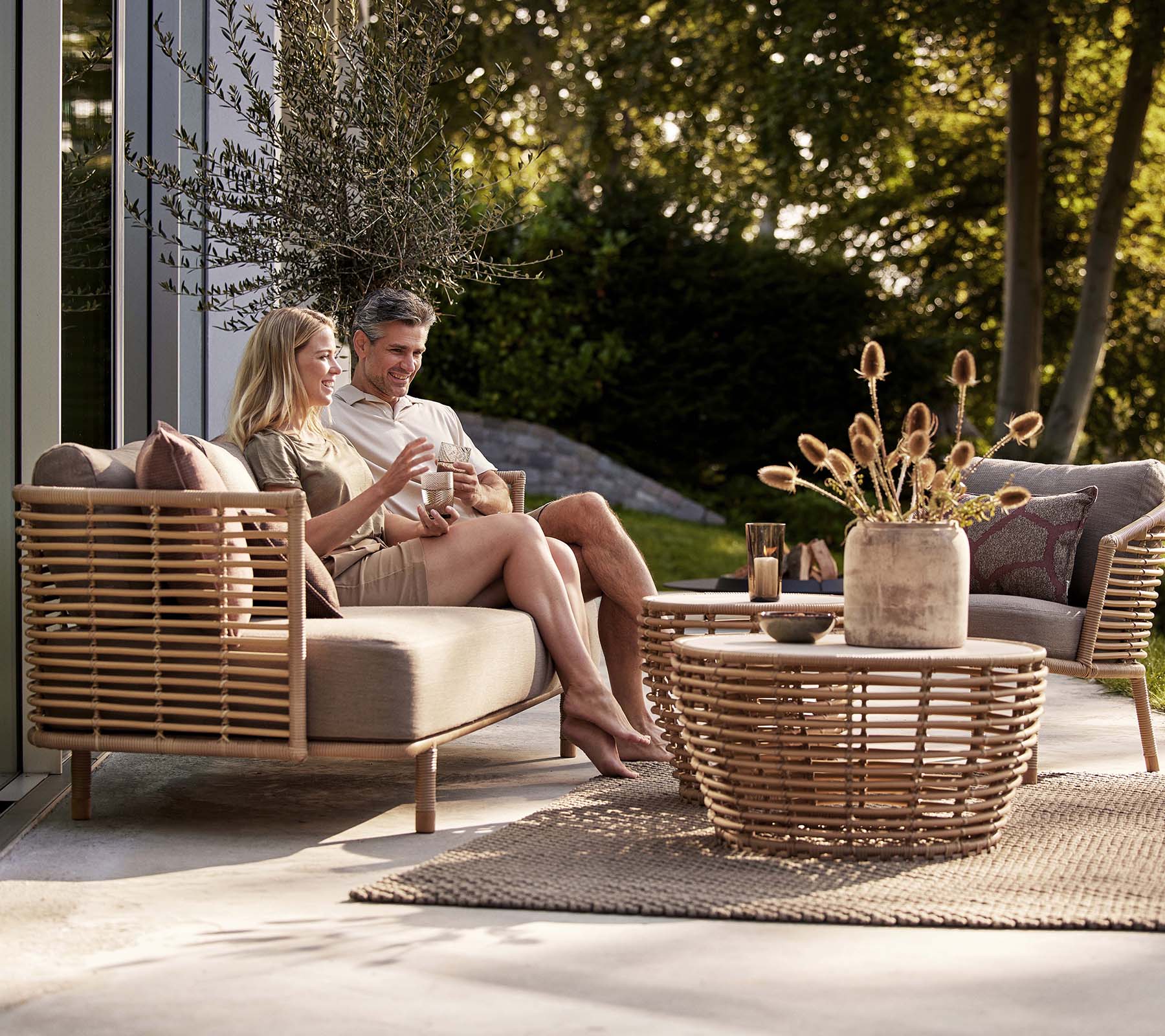 Et par nyder drinks på en Sense-sofa og lounge stol, ledsaget af et stilfuldt kurvekaffebord og en dekorativ vase.