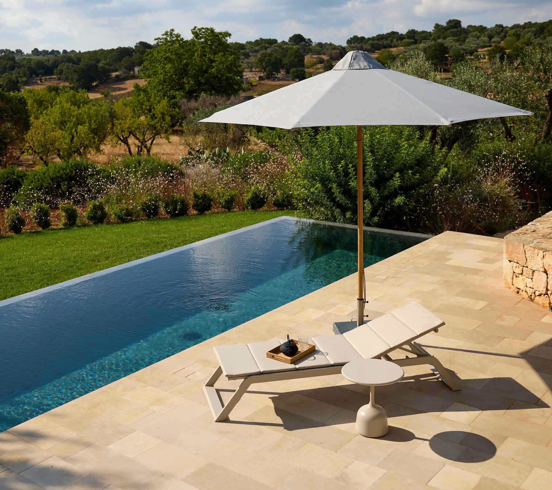 Moderne udendørs loungeområde med en hvid stol, parasol og sidebord ved en pool.