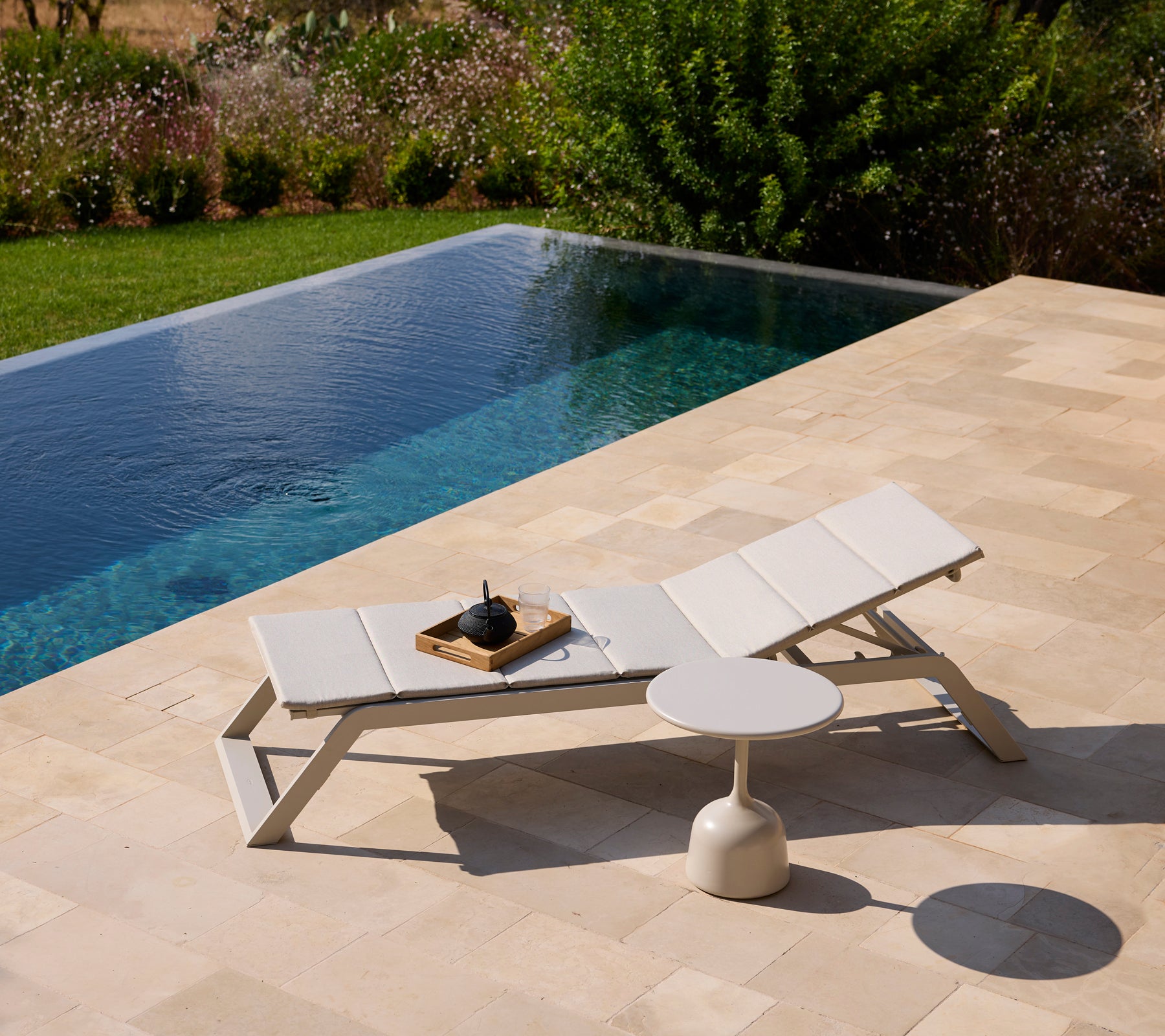 Moderne hvid loungestol og sidebord ved en pool omgivet af grønt område.