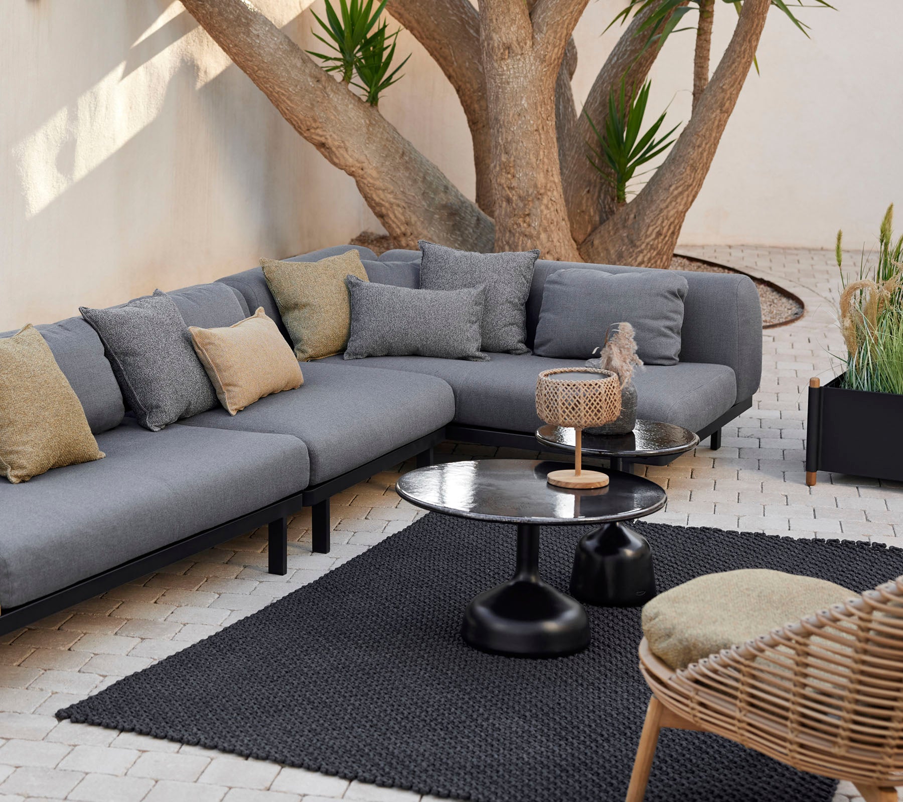 Loungeområde med en grå modulsofa med dekorative puder, et slankt kaffebord og et tekstureret tæppe omgivet af grønt.