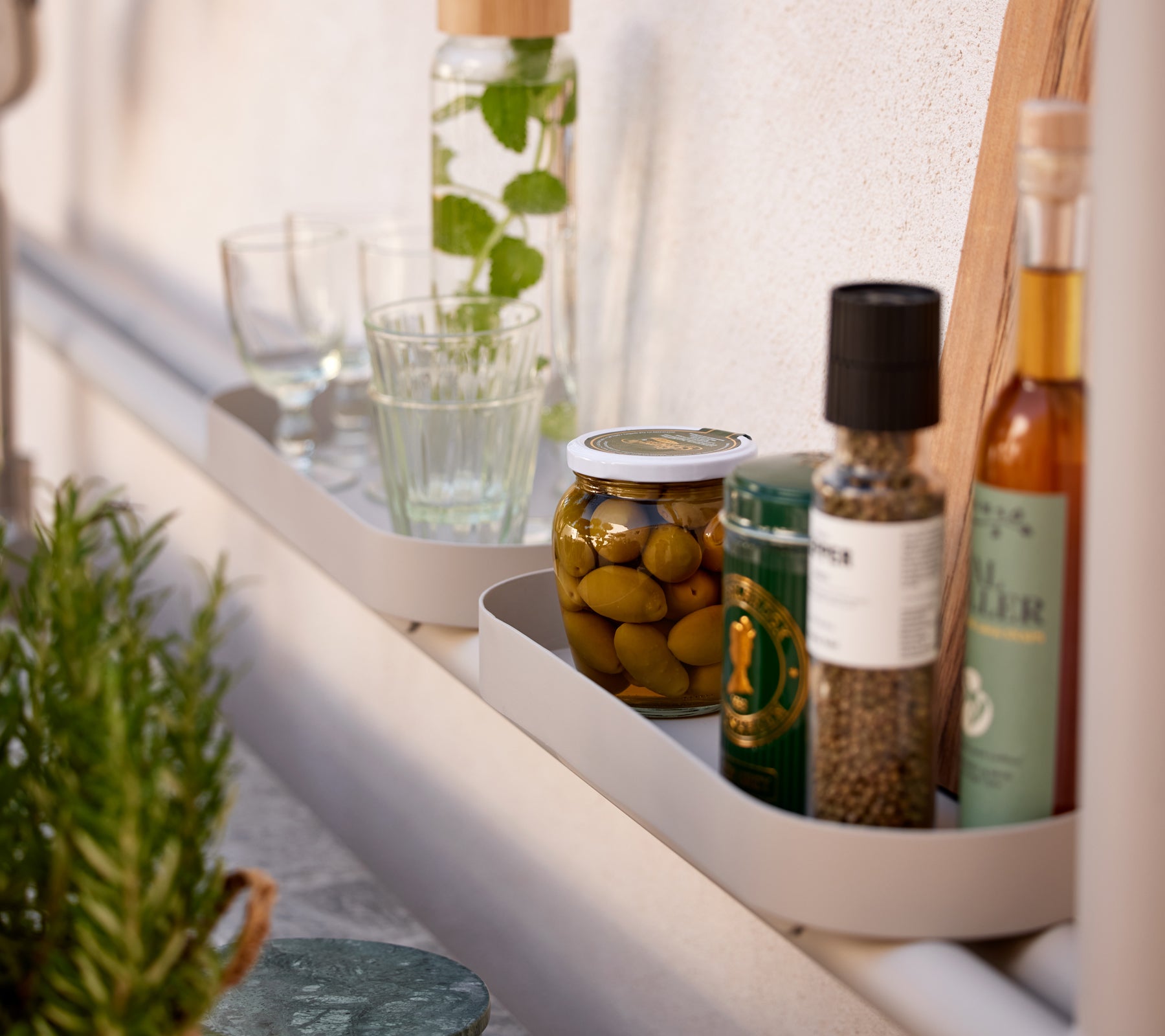En hylde med forskellige køkkenartikler, herunder glas, glas med oliven og flasker med olie og krydderier.
