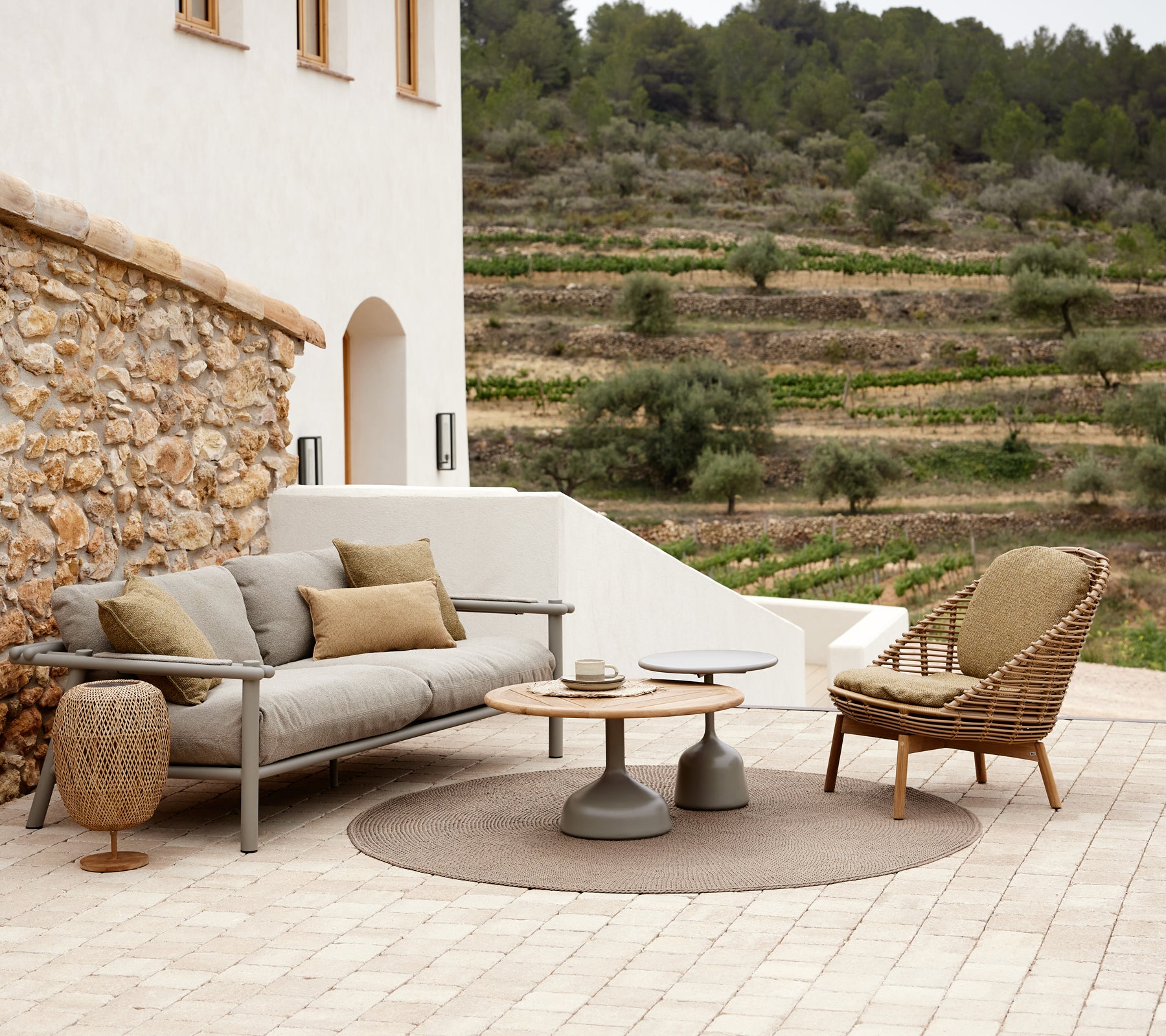 Et stilfuldt loungeområde med en hyggelig sofa med puder, et rundt tæppe, et moderne sidebord og en flettet loungestol, omgivet af grønne planter.