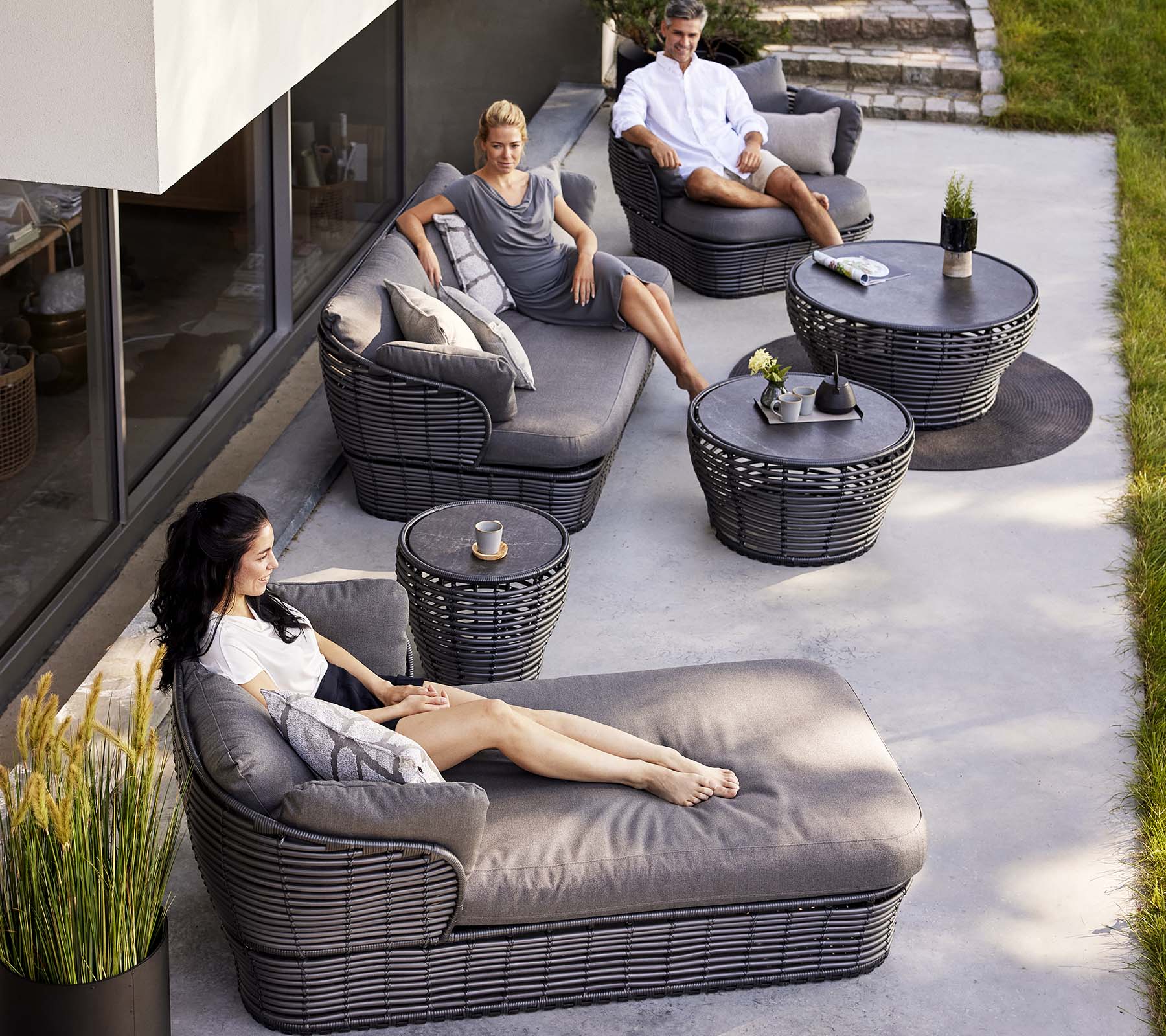 Moderne loungeområde med en kurv-seng, en 3-personers sofa og lounge-stole, suppleret af runde borde og dekorative planter.