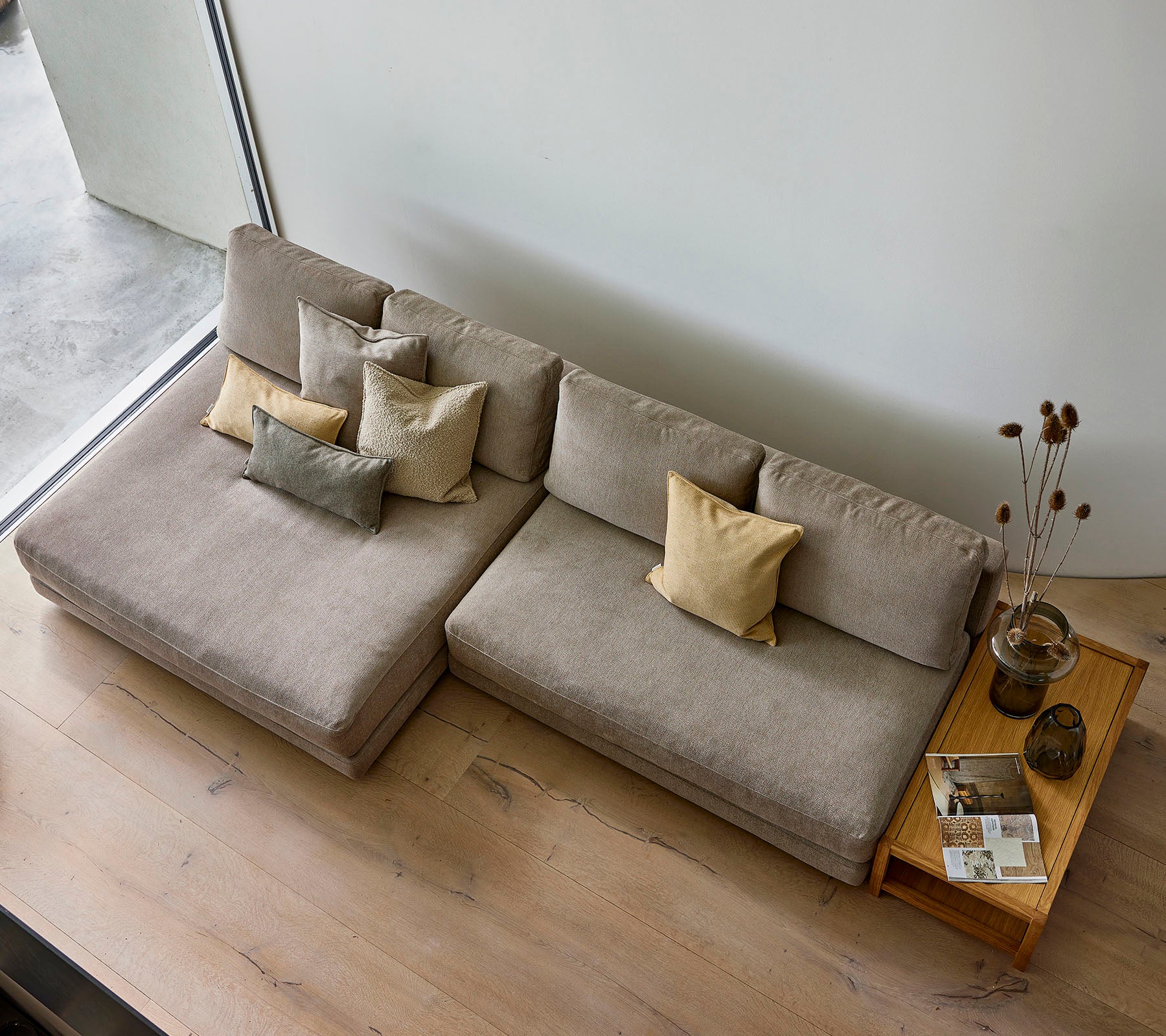 En stilfuld sofa i en neutral tone, prydet med forskellige dekorative puder, ledsaget af et træsidebord med bøger og en vase.