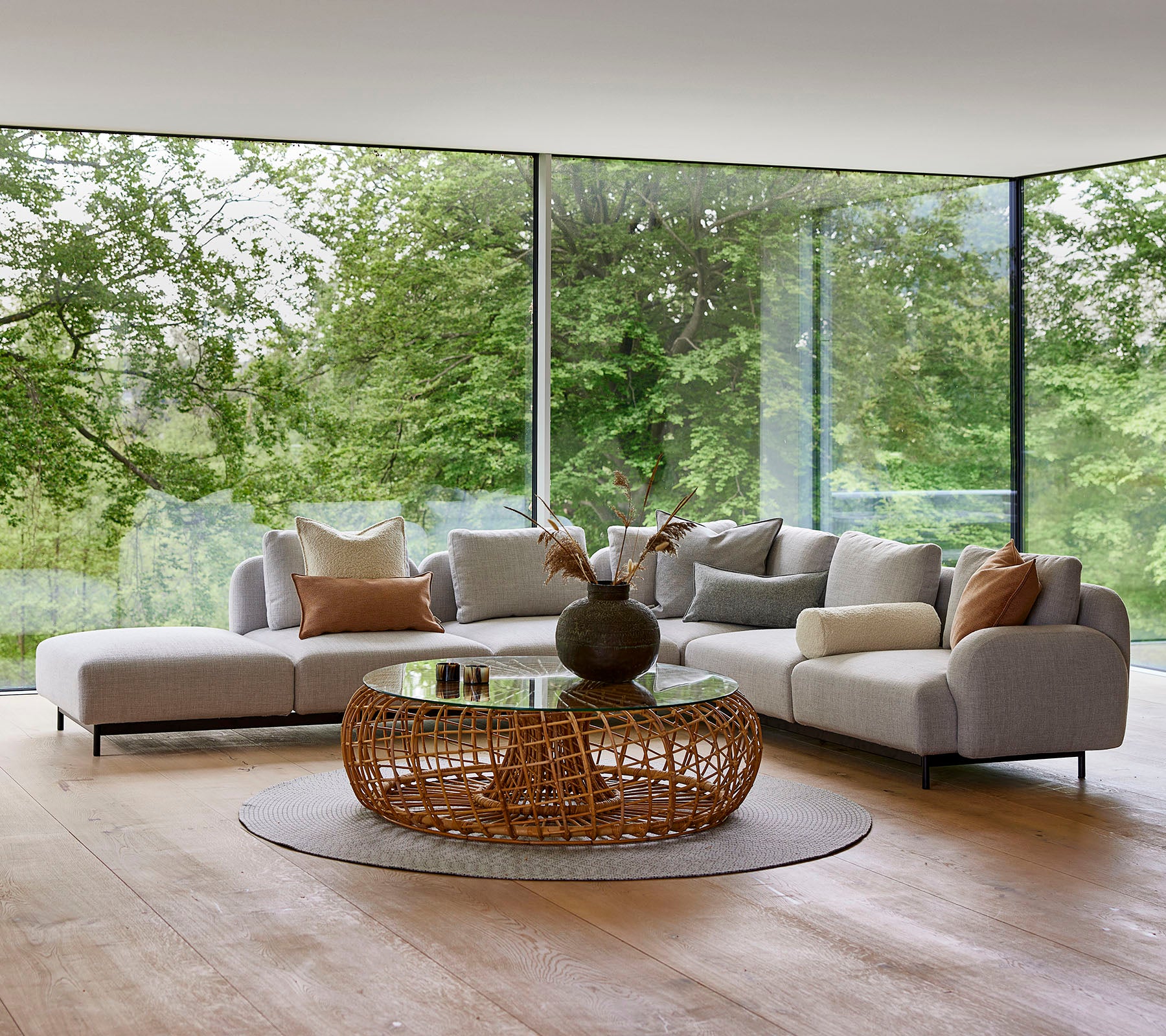 Moderne loungeområde med en modulsofa med dekorative puder, et rundt vævet kaffebord og store glasvinduer, der viser grønt område.
