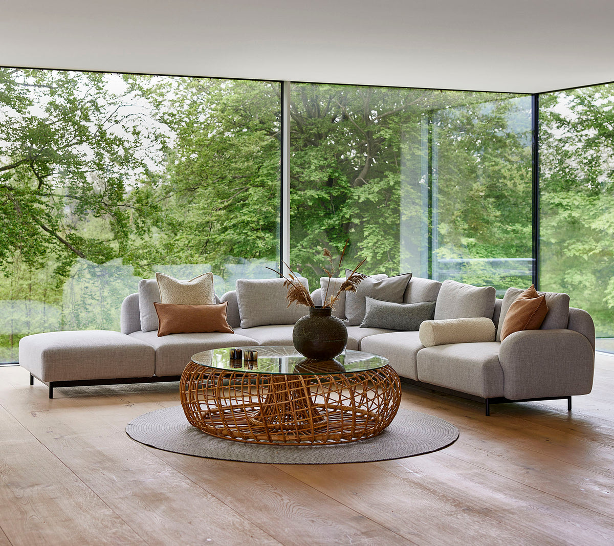 Moderne loungeområde med en modulsofa med dekorative puder, et rundt vævet kaffebord og store glasvinduer, der viser grønt område.