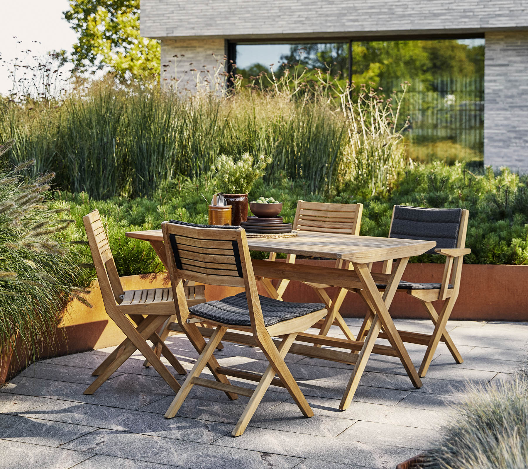 Stilfuldt træbord med matchende stole omgivet af frodig grønne planter og en moderne arkitektonisk baggrund. Perfekt til udendørs sammenkomster.