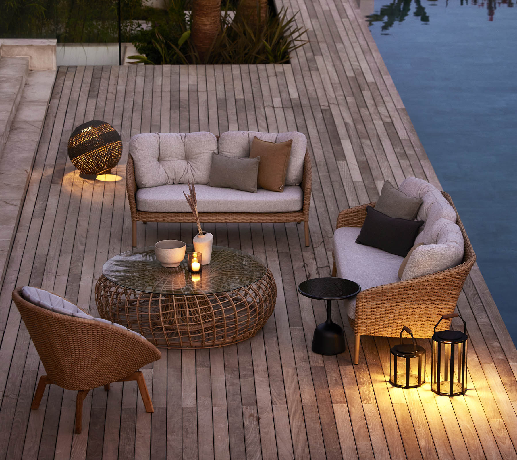 Hygge udendørs siddeområde med vævet møblement, bløde puder og stemningsfuld belysning ved en pool på en træterrasse.